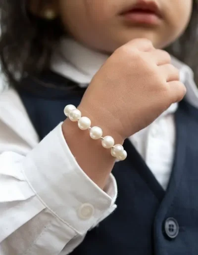 The image shows a child wearing a white pearl bracelet. The focus is on the child's hand and the bracelet, with a blurred background. The pearls are evenly spaced, creating an elegant yet simple look.